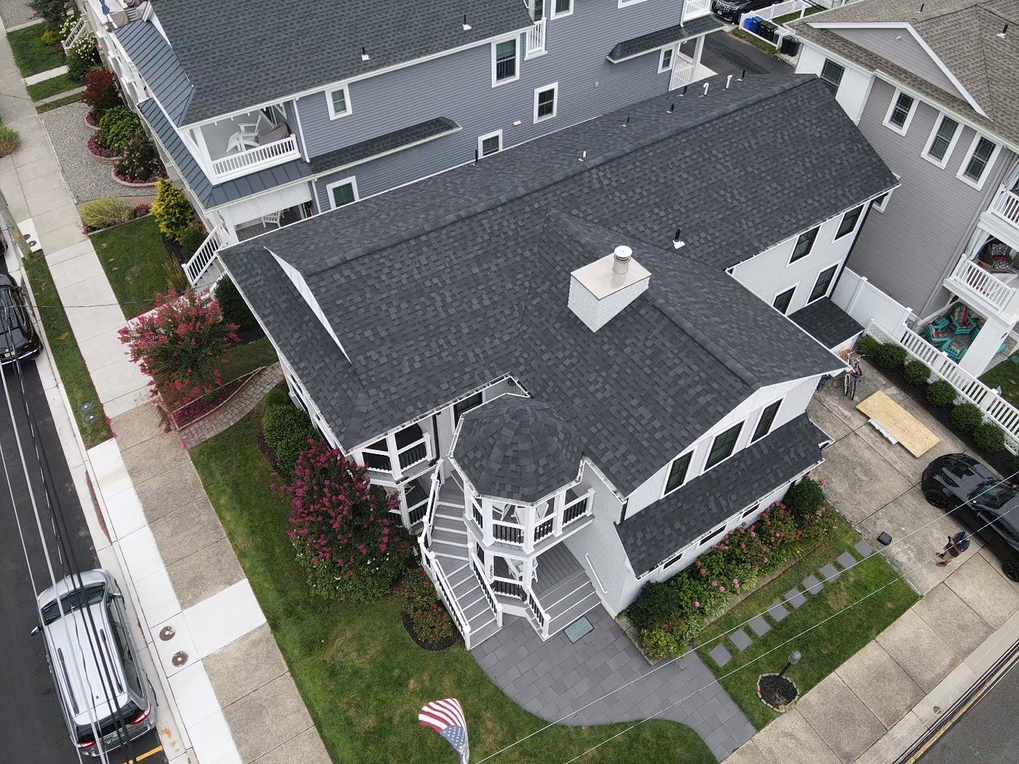 Aerial view of a residential property in Ocean City, NJ, featuring a newly installed dark asphalt shingle roof, landscaped yard with flowering plants, and a driveway with parked vehicles, highlighting Costello Roofing's quality roofing services.